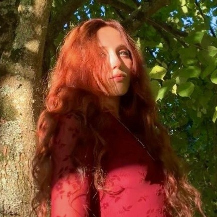 Portrait of Lola Benenati outdoors against a tree, long red hair, wearing a red top.